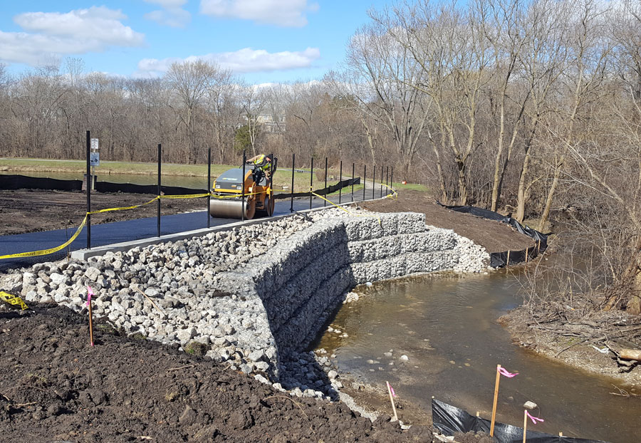 BGPD-Crossings-20190412_100041 A construction site featuring a roller compacting gravel near a newly built stone retaining wall alongside a stream.