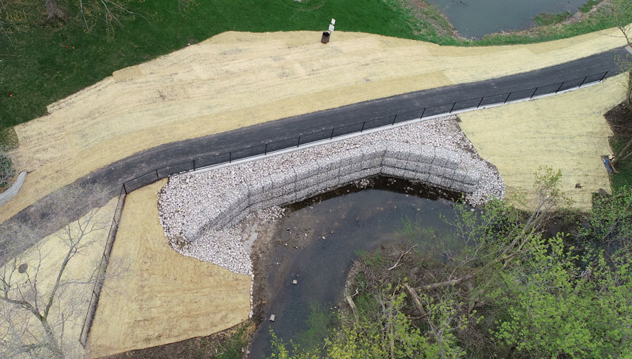 BGPD-Crossings-DJI_0016 Completed erosion control project featuring a stone-lined bank along a stream, with newly paved path and grassed area.