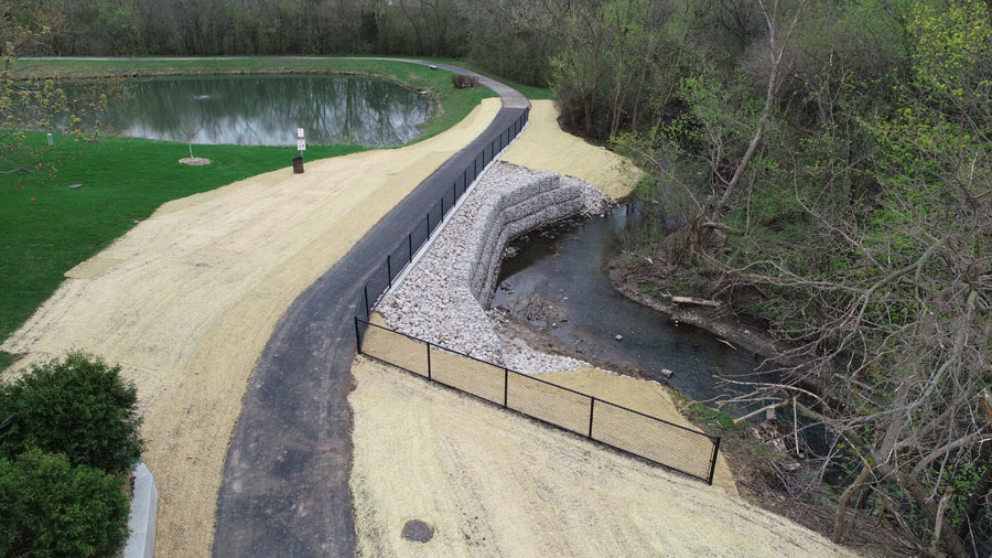 BGPD-Crossings-DJI_0021 View of a newly constructed bank stabilization project along a creek, featuring a gravel retaining wall and a paved walking path.