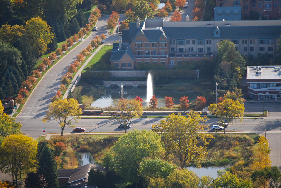 Garlands-DSC_2685-OPT Aerial view of a landscaped park featuring a pond with a fountain, surrounded by autumn trees and adjacent to a residential building and road.