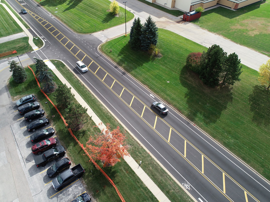 Hook-Orchard-DJI_0529 Aerial view of a newly paved roadway with marked lanes and adjacent parking spaces, surrounded by green grass and trees.