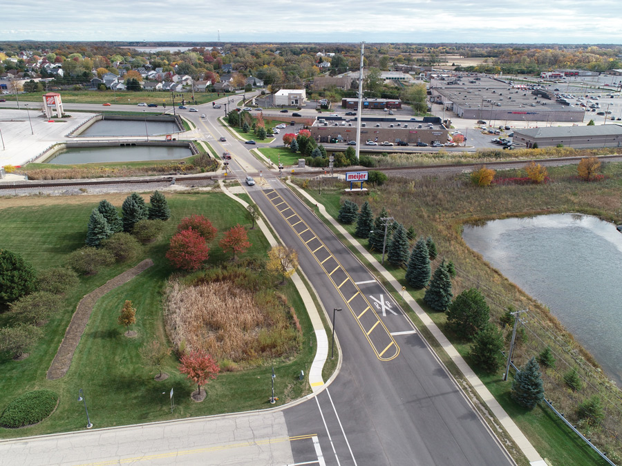 Hook-Orchard-DJI_0540 Aerial view of a newly paved road with marked crosswalks near a retail area, featuring adjacent ponds and landscaped green spaces.