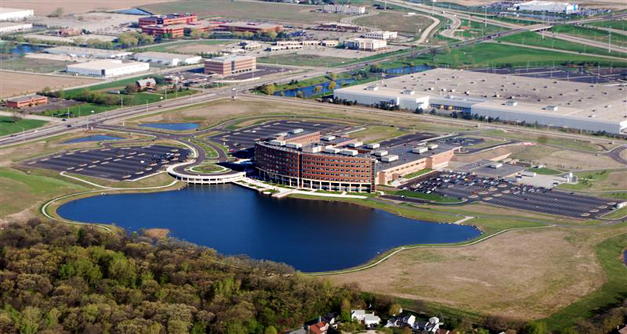 Sherman-Hospital-1 Aerial view of a modern office building by a large pond, surrounded by landscaped grounds and parking areas, with industrial developments nearby.