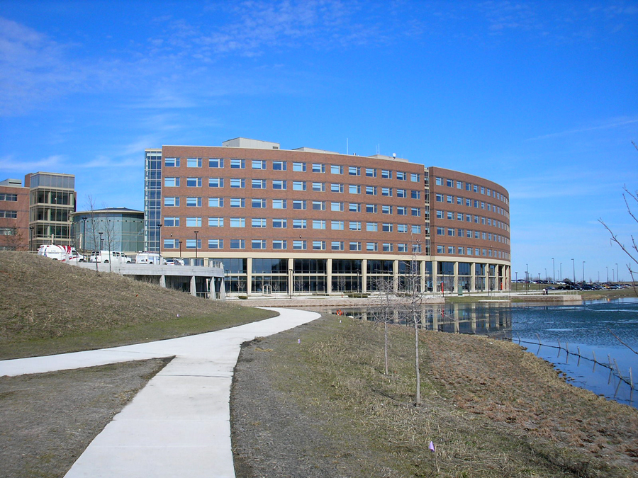 Sherman-Hospital-3 Modern building alongside a paved walkway and water feature, with green space and clear blue sky in the background.
