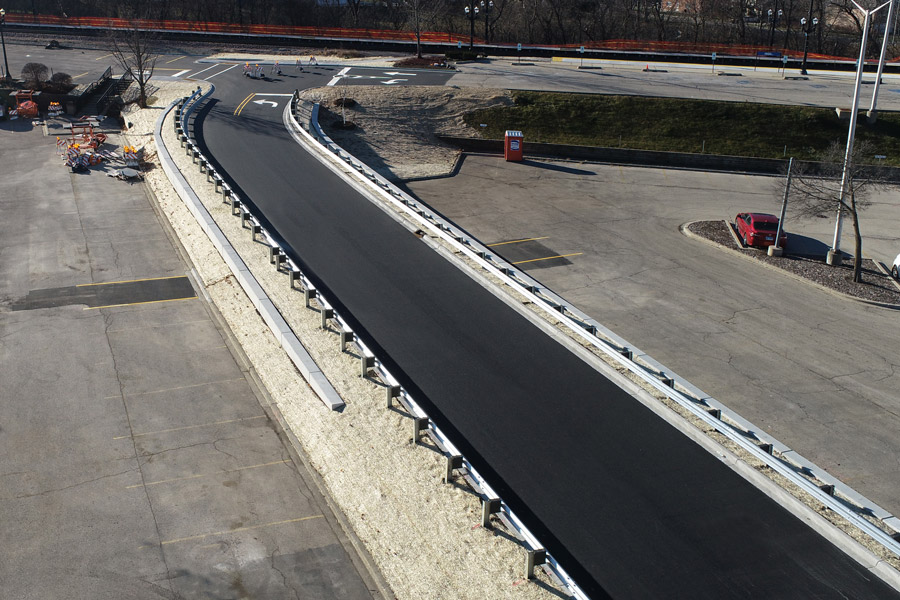 Barr-Metra-DJI_0002 Newly constructed roadway featuring a smooth asphalt surface, protective barriers, and adjacent construction materials in a well-lit area.