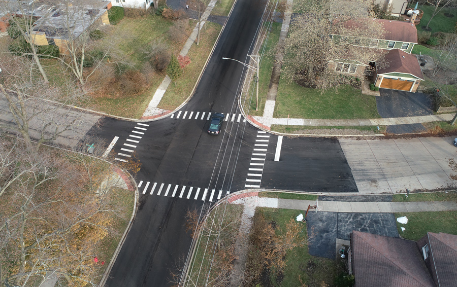 DF-Greenwood-DJI_0015 Aerial view of an intersection featuring newly paved roads and crosswalks, surrounded by residential properties and greenery.