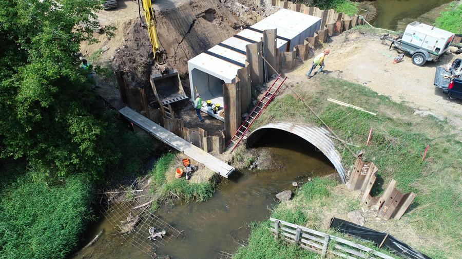 Edwards-DJI_0019-web Construction site showing workers installing concrete culverts at a stream, with heavy machinery and completed bridge nearby.