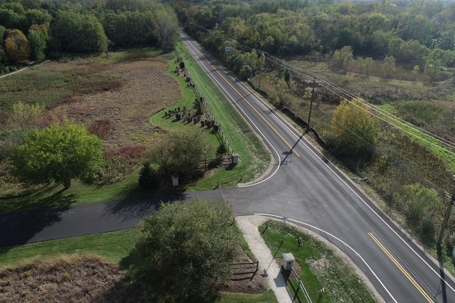 LG-Krueger-DJI_0106 Aerial view of a winding road next to green fields and trees, showcasing a blend of infrastructure and natural landscape.