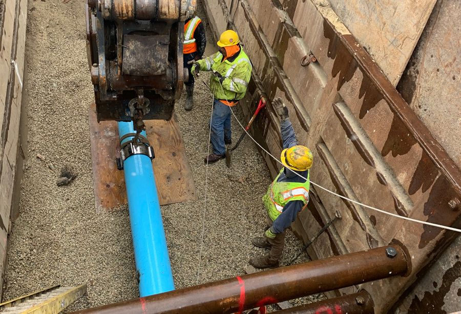 Midland-LCPW-Installing-pipe-in-casing Workers installing a large blue pipeline in a trench, using machinery and safety gear, with gravel surrounding the pipe.