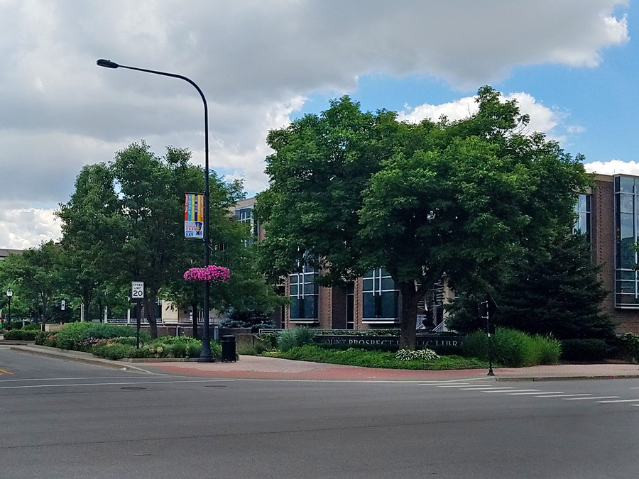 20200713_121654_Web Street view displaying trees, flower baskets, and a municipal building with large windows, set against a partly cloudy sky.
