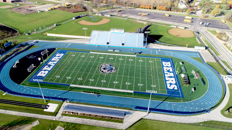 DJI_0004-Web Aerial view of a modern athletic stadium with a blue track, football field, and surrounding green space, showcasing facility upgrades.
