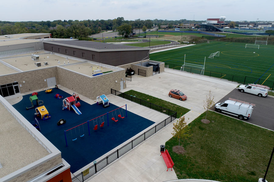 DJI_0141-Web Aerial view of a playground with equipment, adjacent to a sports field and municipal buildings, showcasing infrastructure and recreational areas.