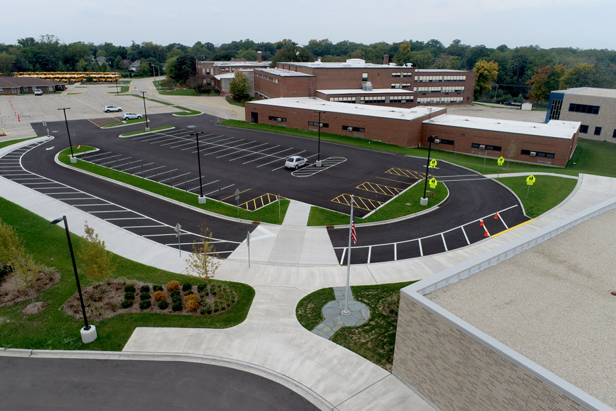 DJI_0142-Web Aerial view of a school parking lot featuring newly paved lanes, marked spaces, and landscaped areas, with nearby buildings visible.