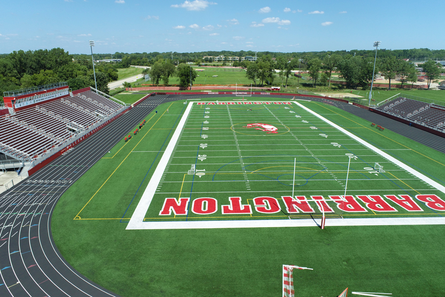BarrHS-DJI_0001-web Aerial view of a sports field with a red and white track, surrounded by green trees and a stadium with seating.