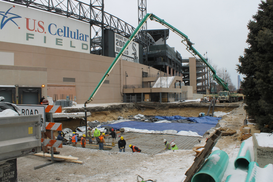 US-Cellular-DJI_0029_web Construction site with workers pouring concrete into a foundation pit, surrounded by construction equipment and safety barriers near a stadium.