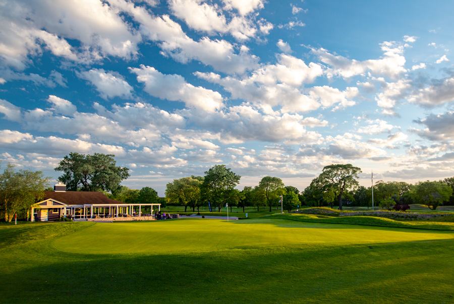 SVGC-9thGreen.Clubhouse.0067-web A well-maintained golf course with a vibrant green lawn, surrounded by trees and a clubhouse under a blue sky with scattered clouds.