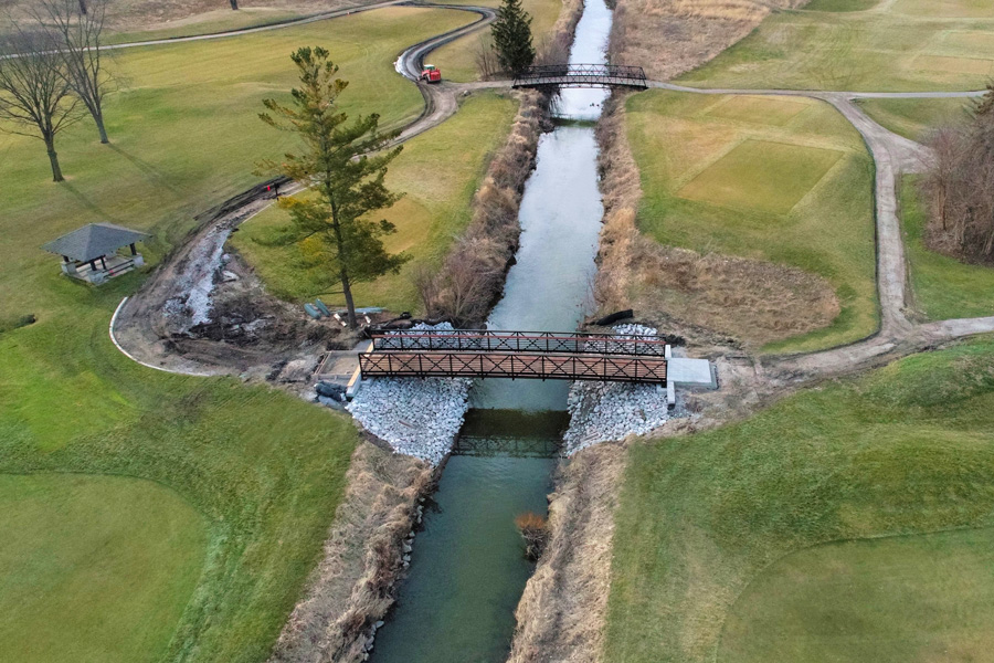SVGC-Bridge_web Aerial view of a new metal pedestrian bridge crossing a stream, surrounded by landscaped areas and a golf course.