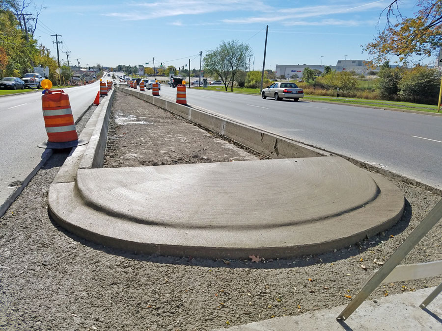 Mellody-VH-Rds_120218_web Curved concrete curb under construction along a busy street, surrounded by traffic cones and gravel, showcasing municipal infrastructure work.