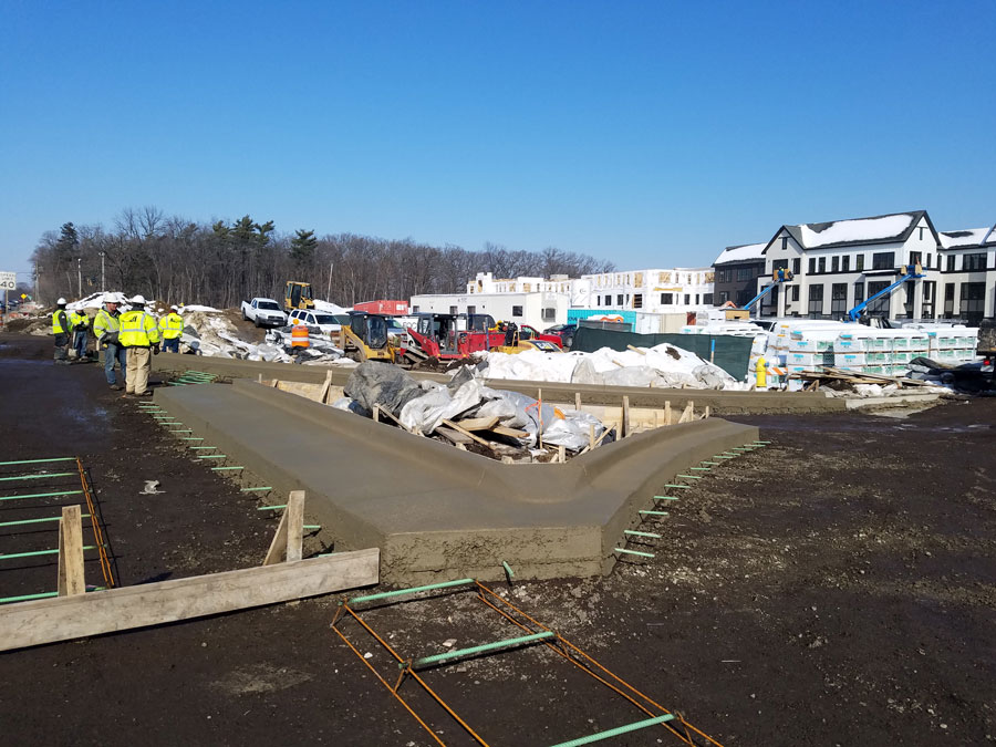 Mellody-VH-Rds_132310_web Construction site with workers pouring concrete for a roadway, surrounded by equipment and partially completed buildings.