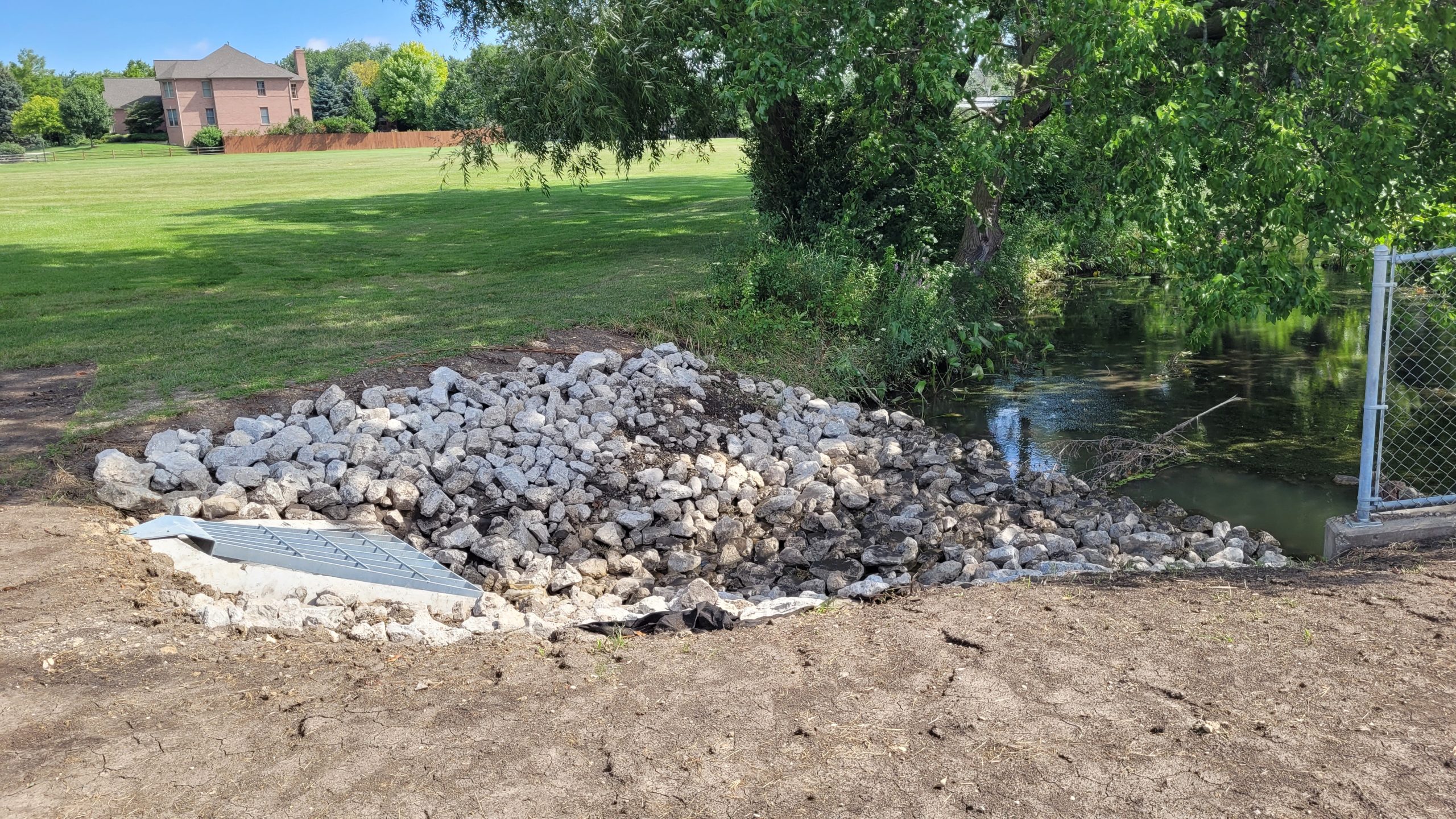 20220727_101846 A pile of rocks placed as erosion control along a water's edge, with a grassy area and a residential building in the background.