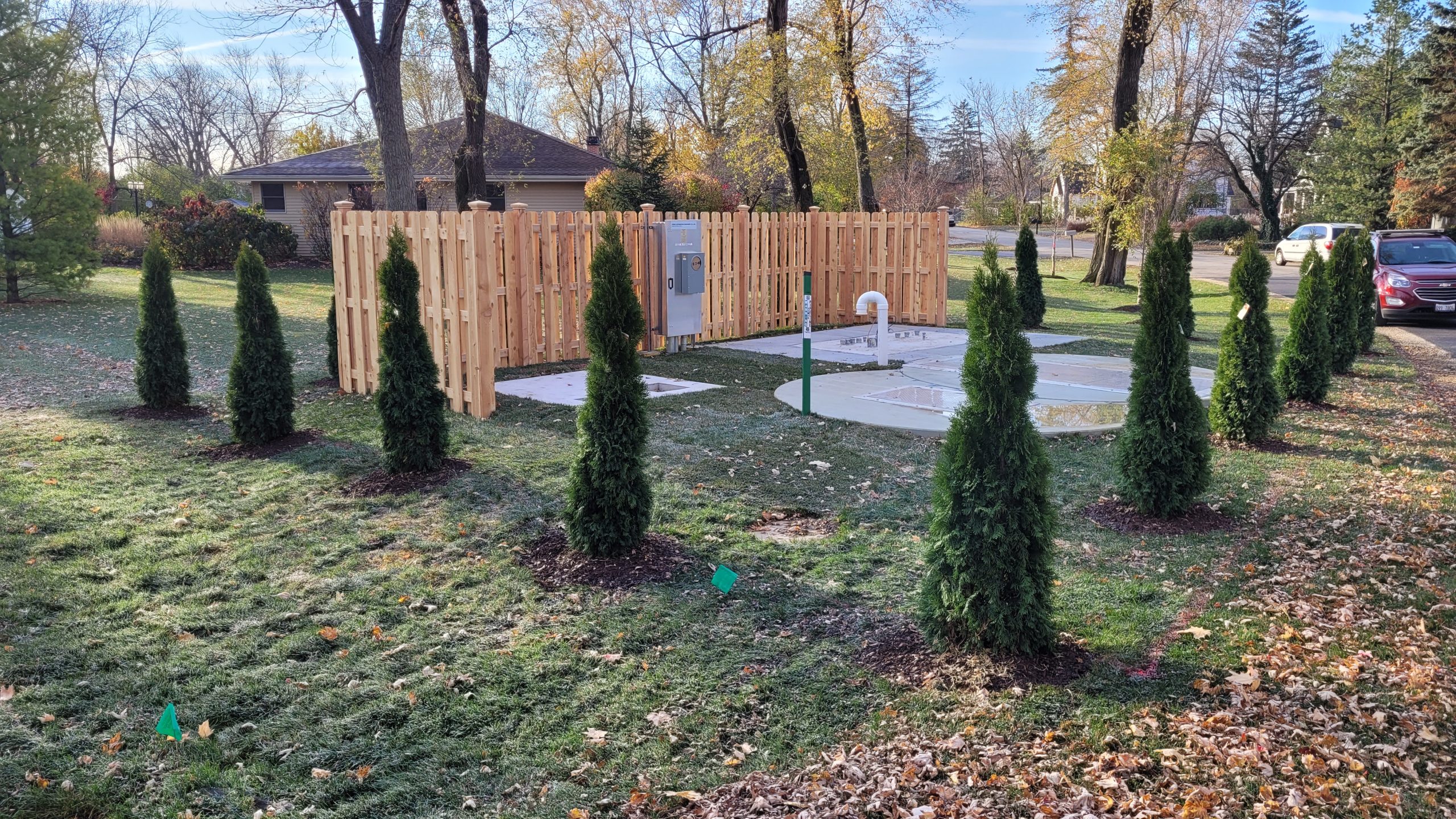 20221114_081844 Landscaped area with evergreen trees surrounding a fenced utility facility, featuring concrete pads and municipal infrastructure elements.
