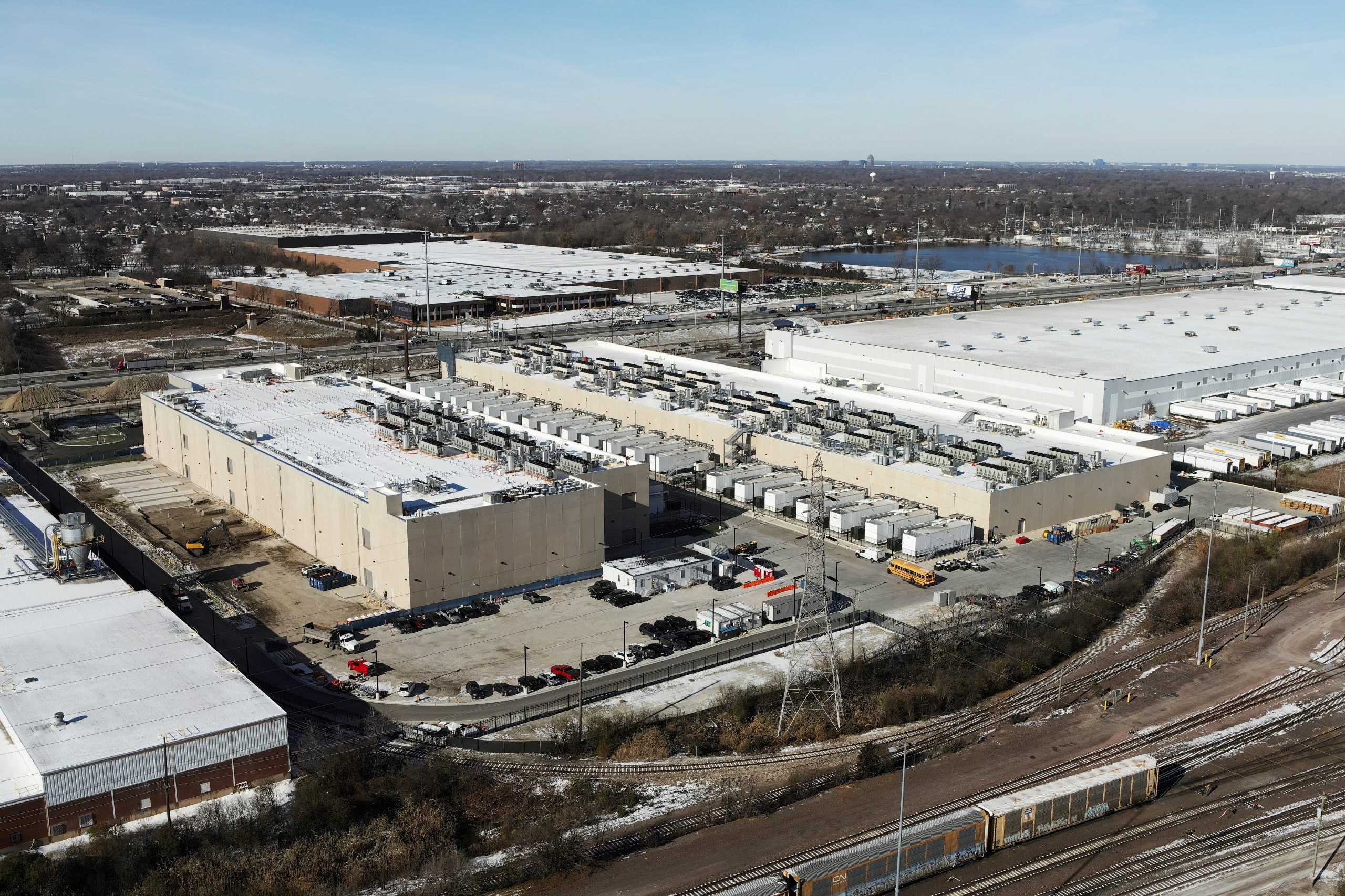 DJI_20231128104954_0001 Copy Aerial view of a large industrial facility with multiple loading docks, surrounded by parking areas and rail tracks.