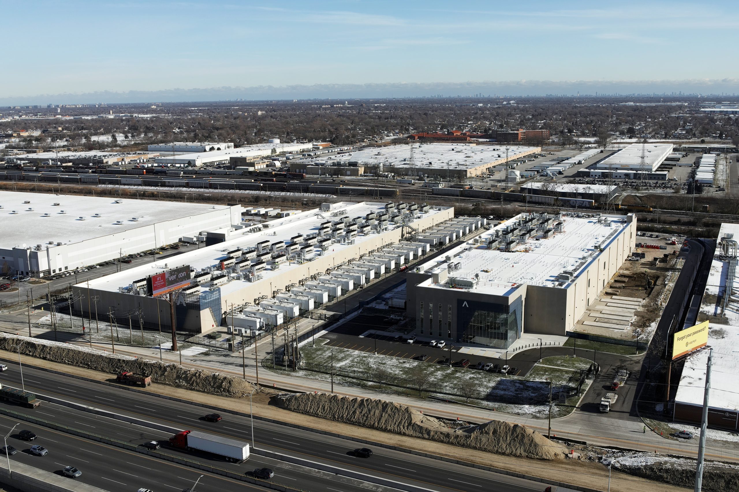 DJI_20231128105229_0004 Copy Aerial view of a large industrial facility with multiple loading docks, surrounded by construction and infrastructure, next to a highway.
