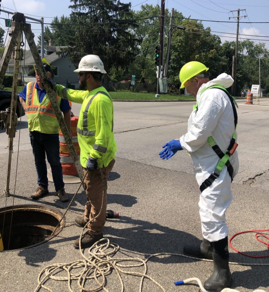 GHA Engineer Technician John Bolton stands next to an open manhole, wearing safety equipment, preparing to perform a manhole inspection.