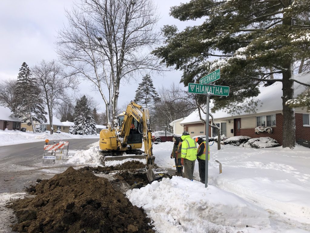 Image of utility work being performed. Three workers in high visibility vests performing utility work with an excavator at the intersection of Bernard and Hiwatha Drives in Buffalo Grove, Illinois.