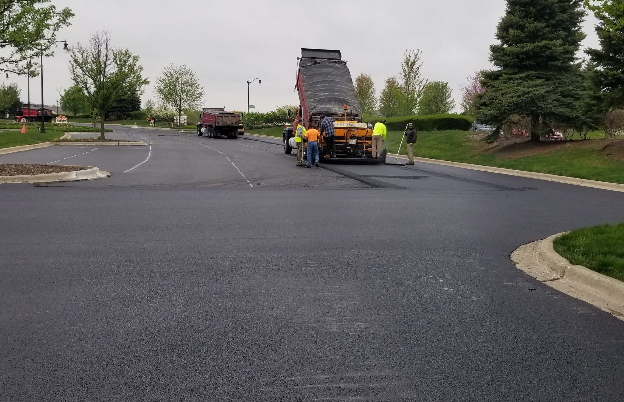 Construction crew paving a new asphalt surface, with machinery and equipment visible in a commercial parking area.