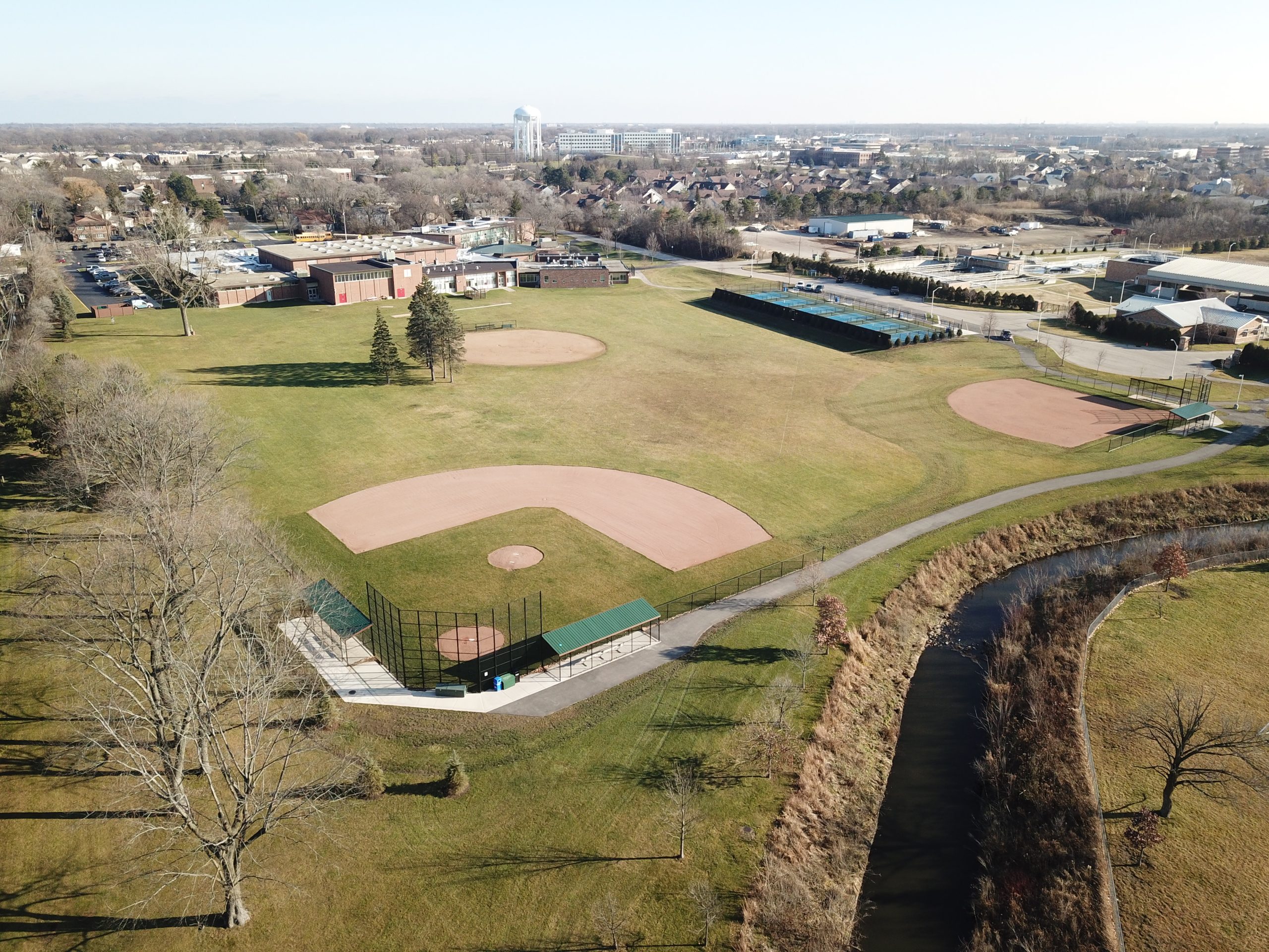 DCIM100MEDIADJI_0001.JPG Aerial view of a baseball field and green park space, featuring manicured grass, pathways, and surrounding structures in an urban setting.