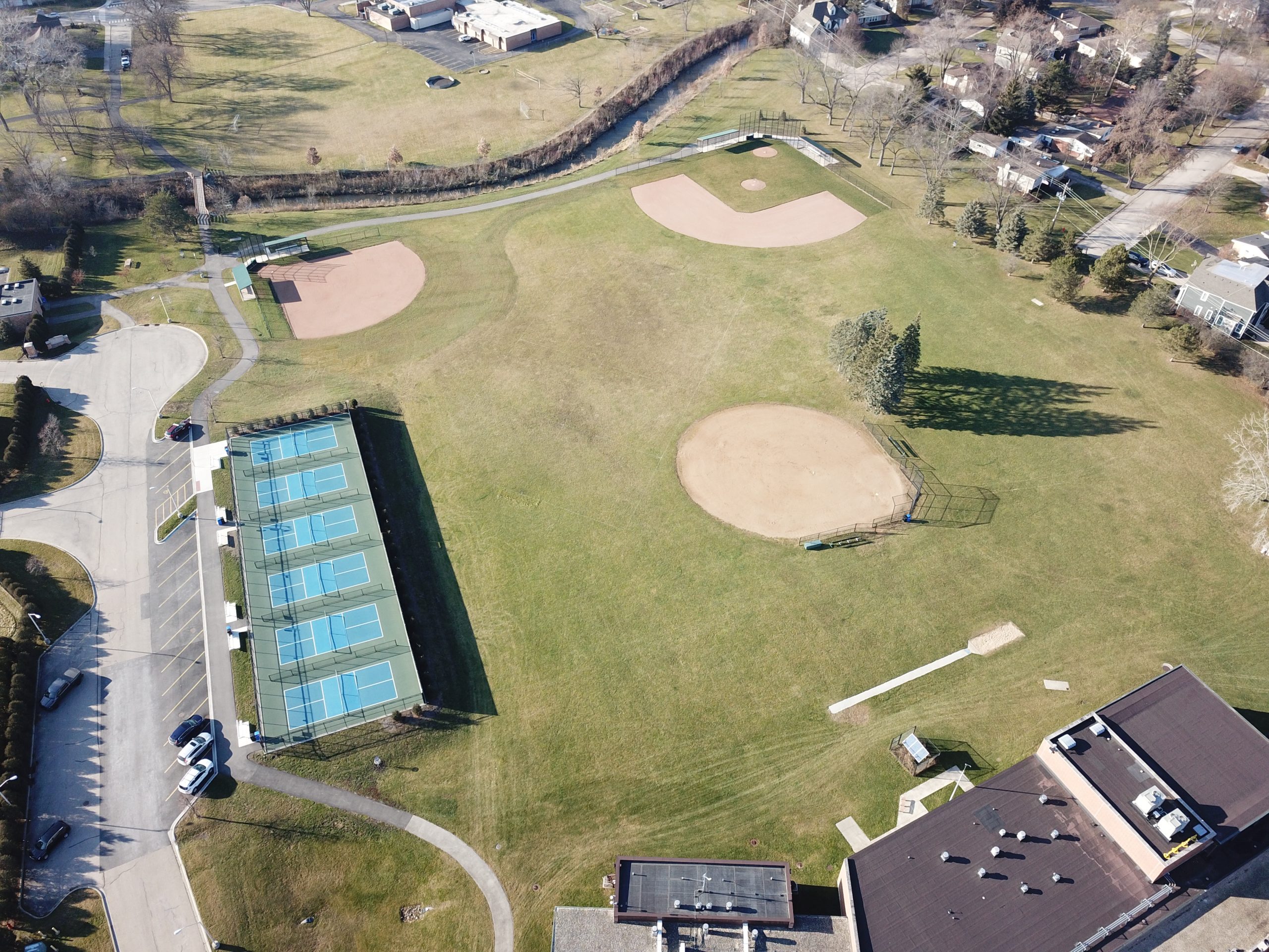 DCIM100MEDIADJI_0059.JPG Aerial view of a public park featuring tennis courts, baseball diamonds, green fields, and surrounding residential areas.