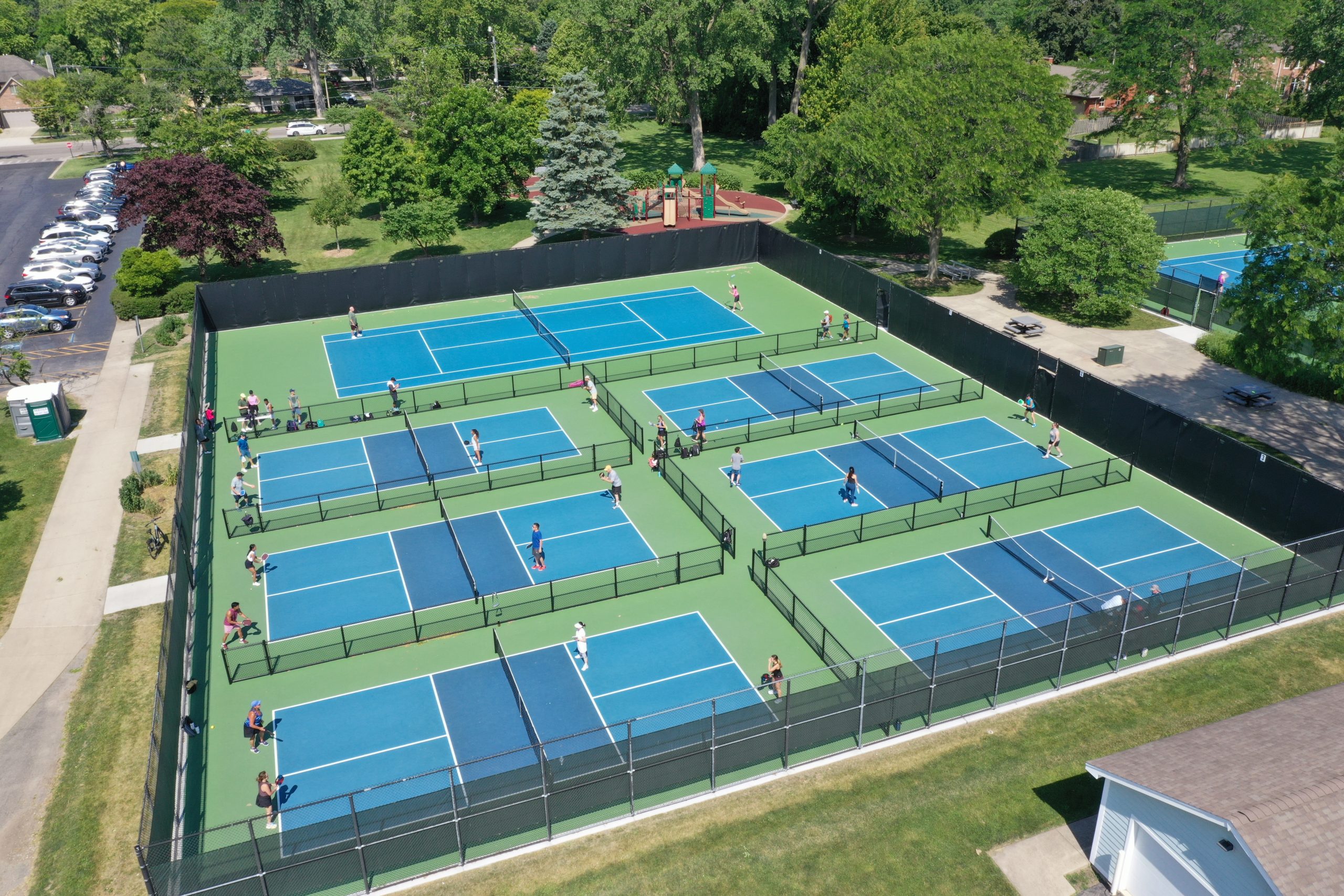 default Aerial view of multiple tennis courts in a park, with players engaged in matches surrounded by greenery and recreational facilities.