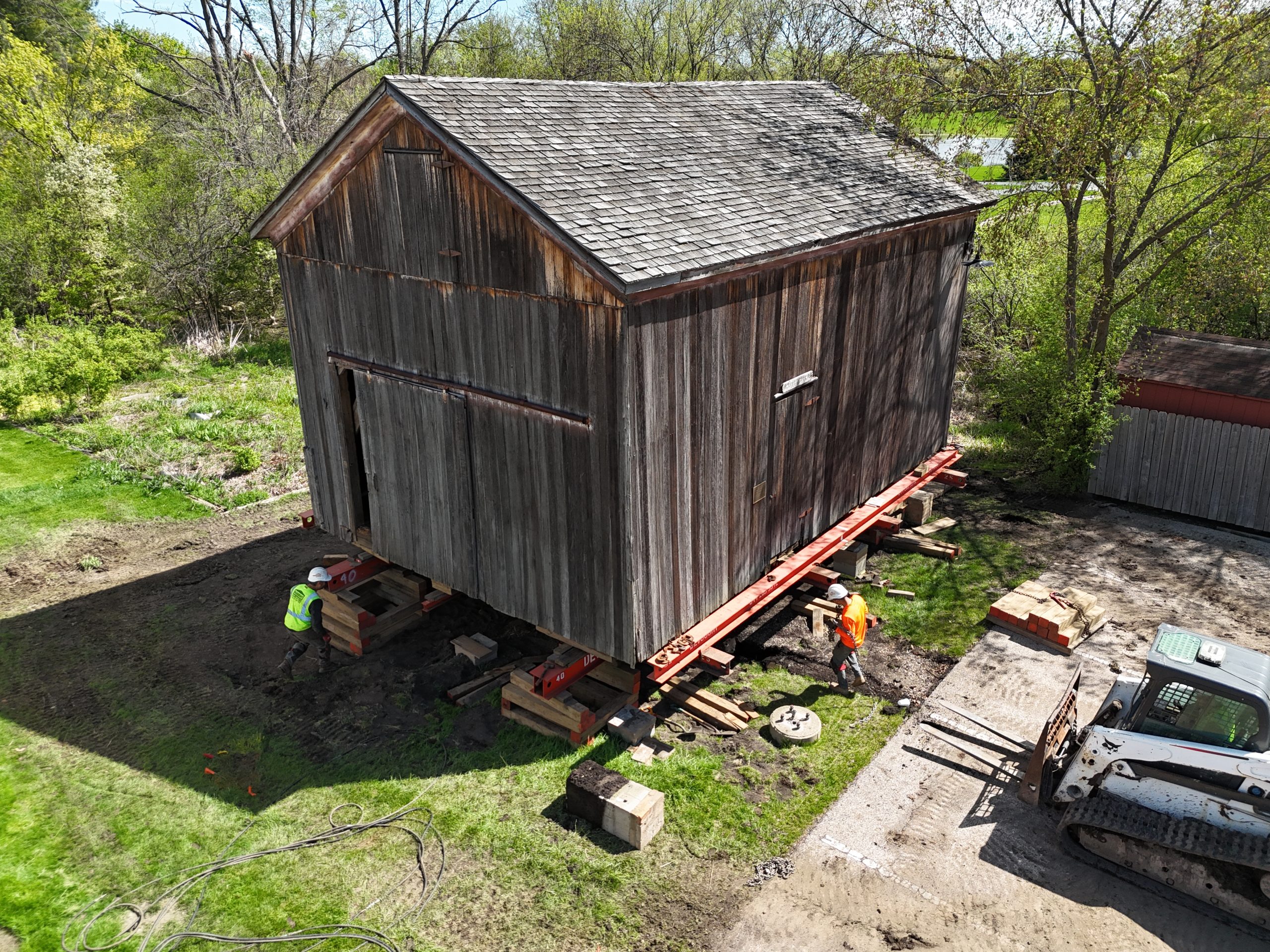 default A wooden structure being relocated on supports, with workers and machinery present in a green outdoor setting.