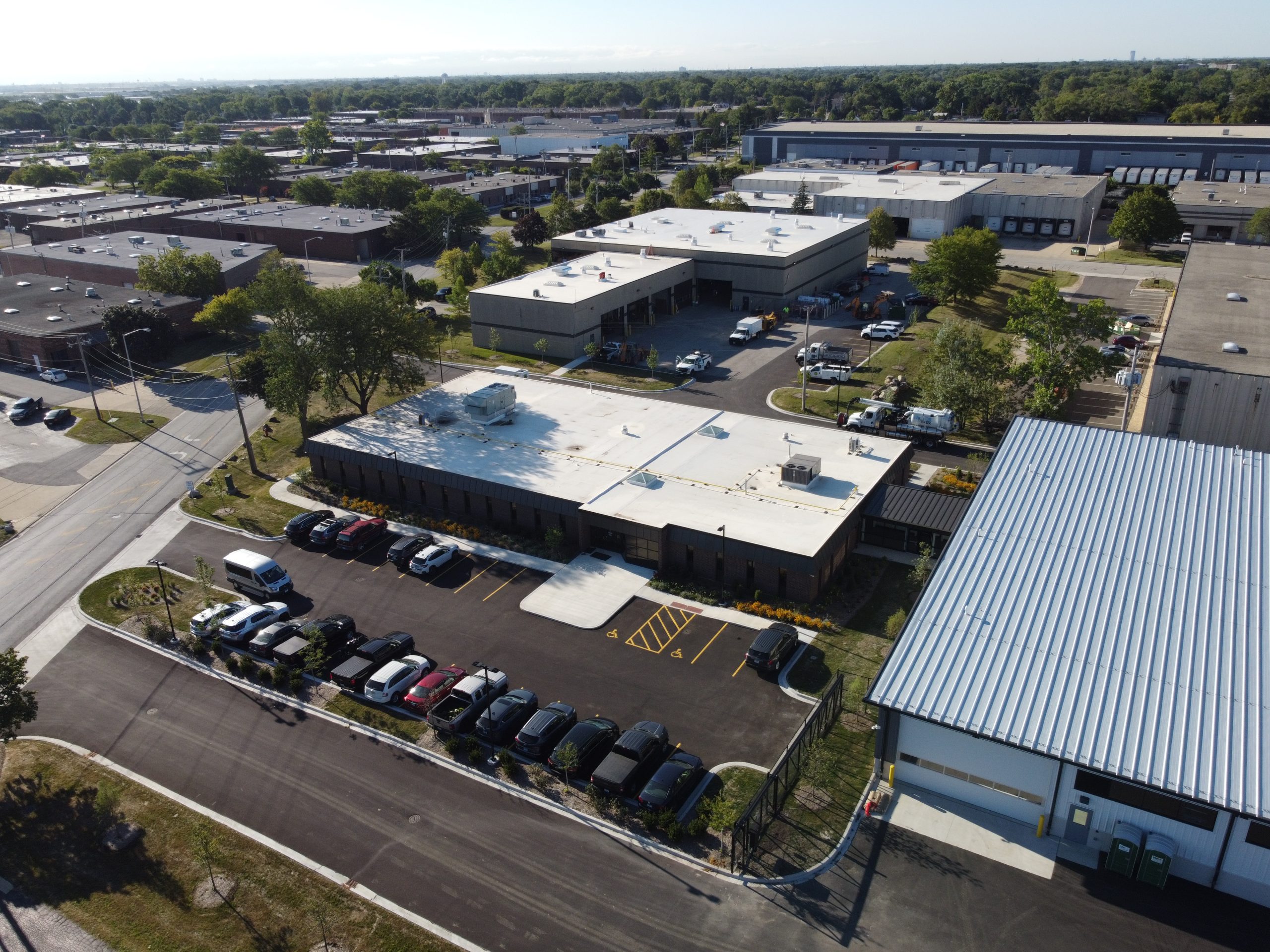 DCIM101MEDIADJI_0211.JPG Aerial view of a commercial building with a parking lot filled with vehicles, surrounded by industrial and municipal structures.