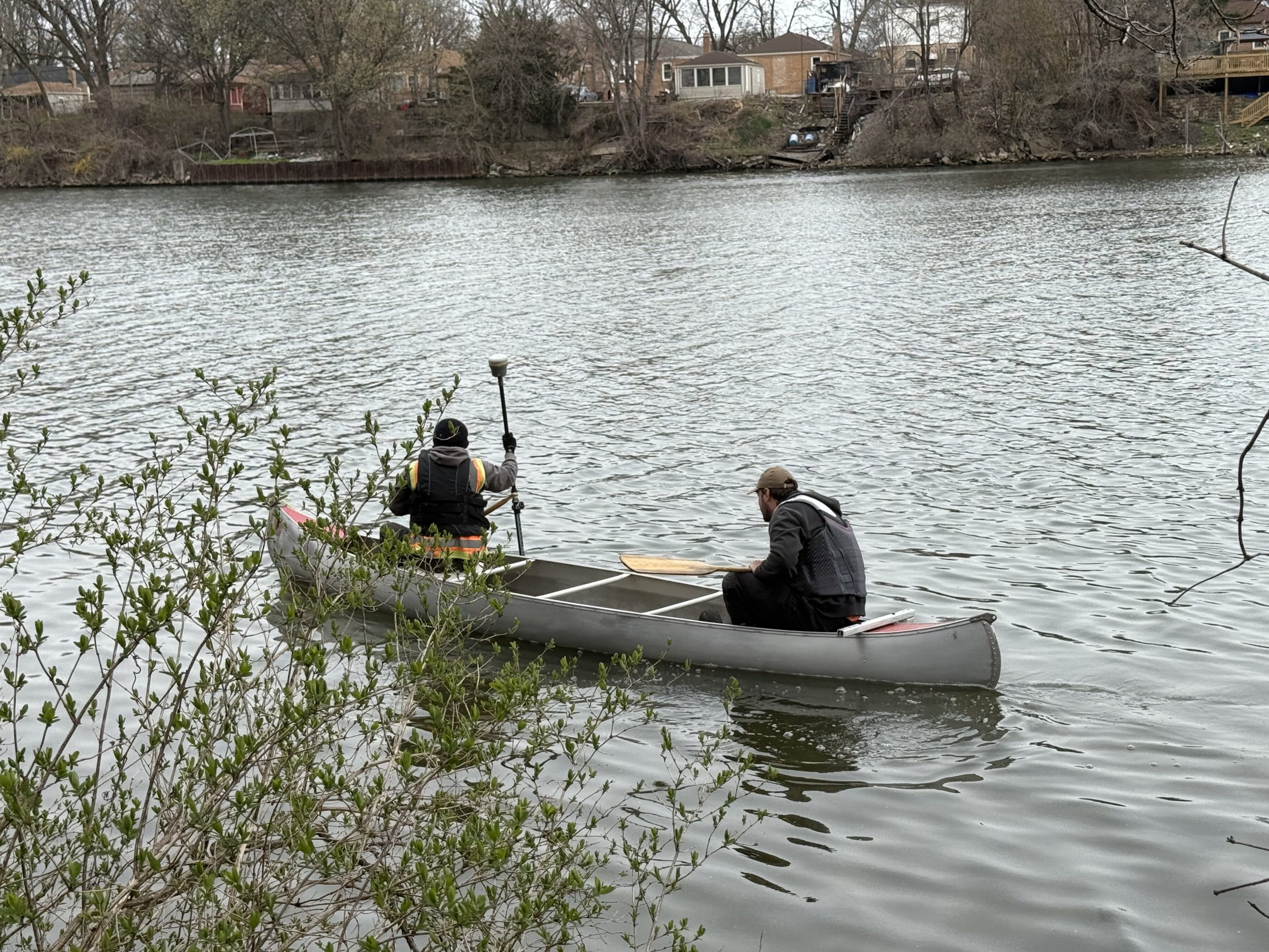 IMG_0115 Two individuals in a canoe paddling on a river, surrounded by greenery and residential homes in the background.