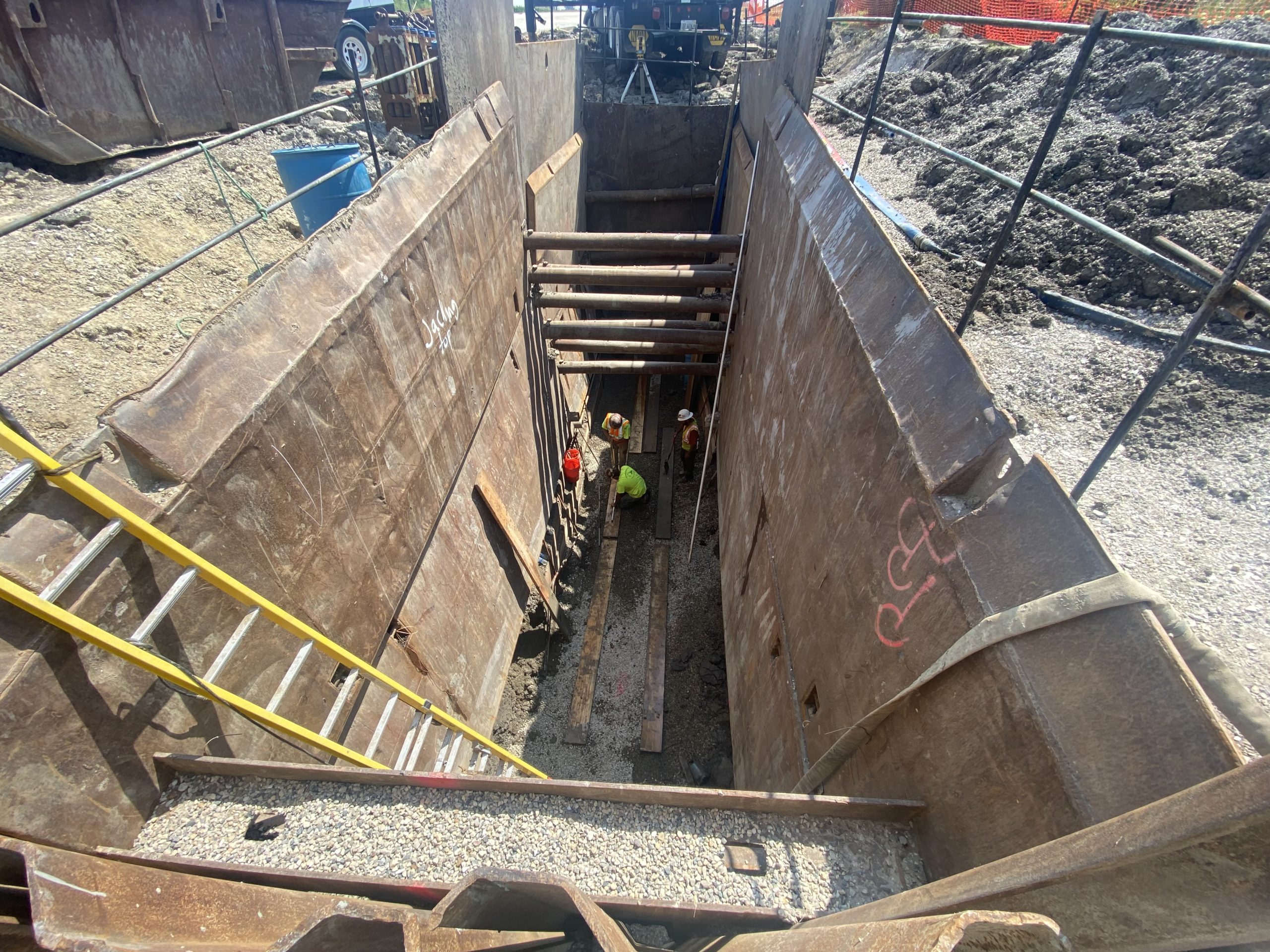 IMG_8956 Construction workers inside a deep excavation site, featuring support beams and safety equipment, surrounded by soil and construction materials.