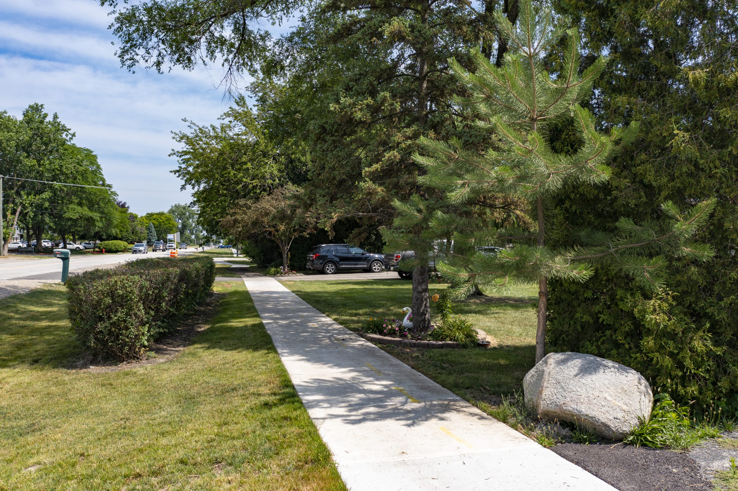 Wolf Rd-4 A well-maintained sidewalk lined with trees and shrubs, leading to a landscaped area in a suburban setting.