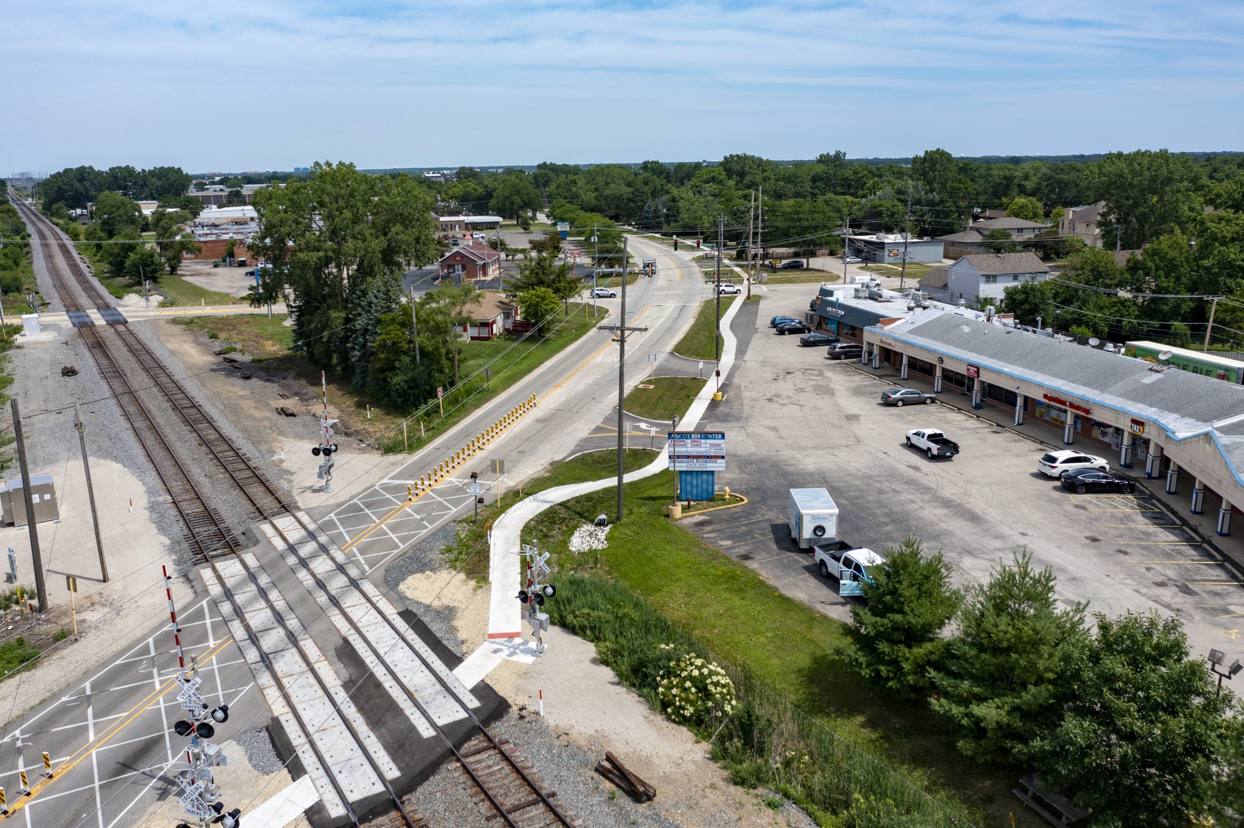 Wolf Rd-5 Aerial view of a road crossing railway tracks with adjacent commercial buildings and parking areas, surrounded by greenery.