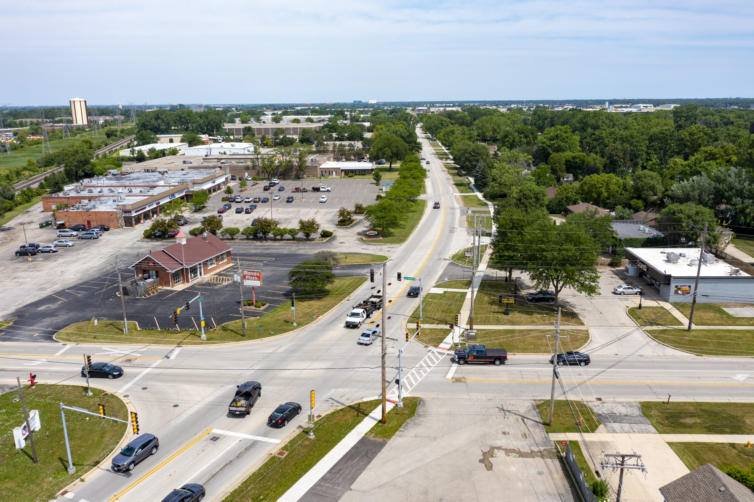 Wolf Rd-6 Aerial view of a busy intersection featuring roads, parking lots, and surrounding green areas with trees and buildings in the background.
