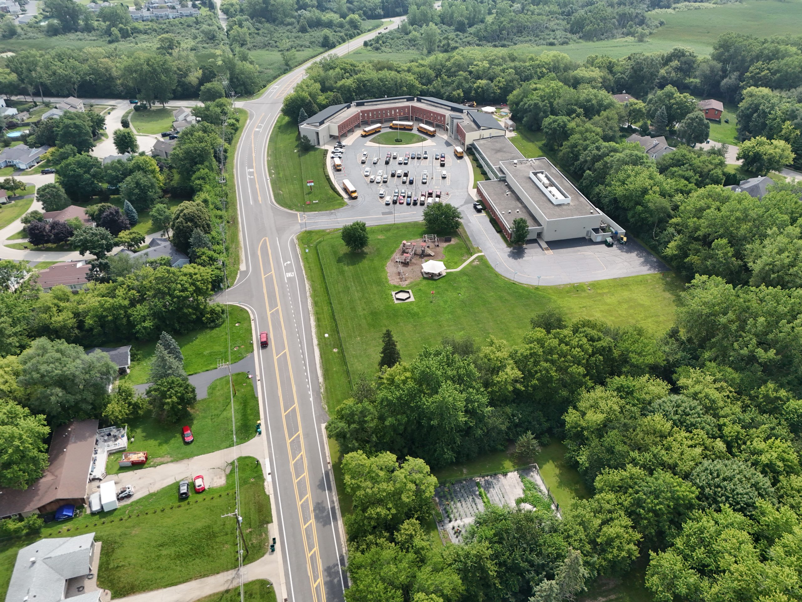 default Aerial view of a municipal area featuring a school, parking lot, and surrounding green space, bordered by a road and trees.