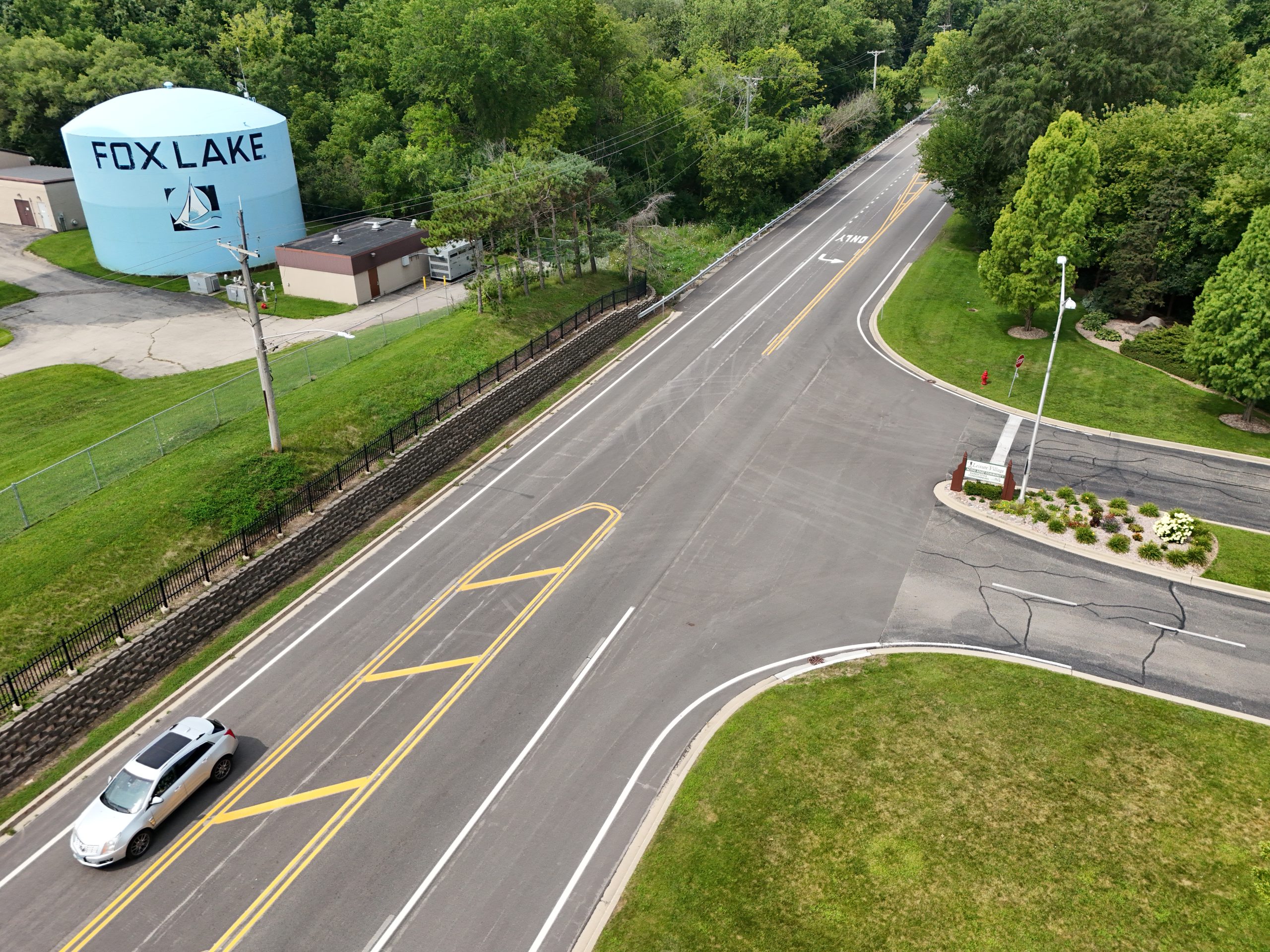 default Aerial view of a roadway intersection with a parked car, surrounded by lush greenery and the Fox Lake water tower in the background.
