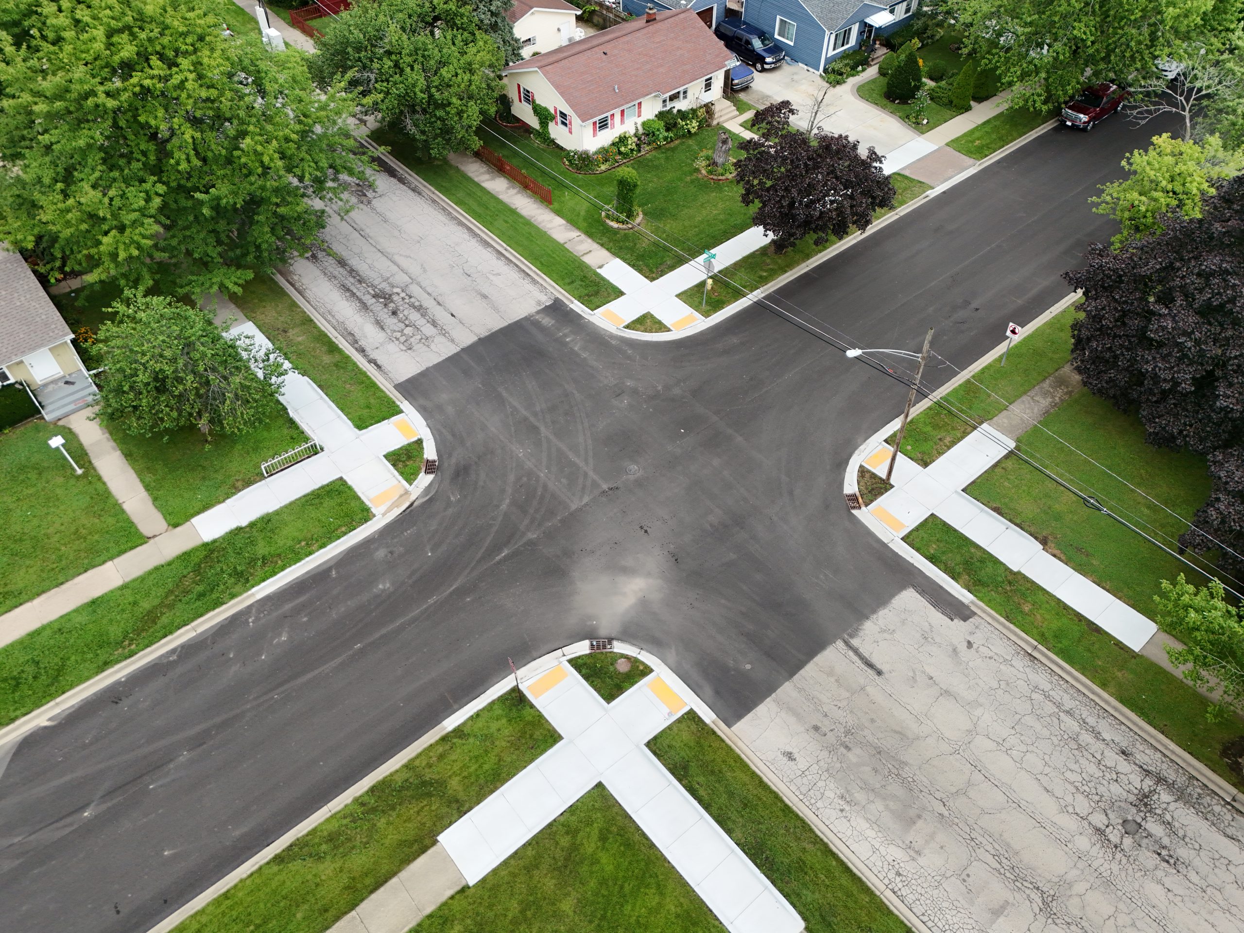 default Aerial view of a residential street intersection, featuring new asphalt, sidewalks, and landscaped lawns surrounding homes.