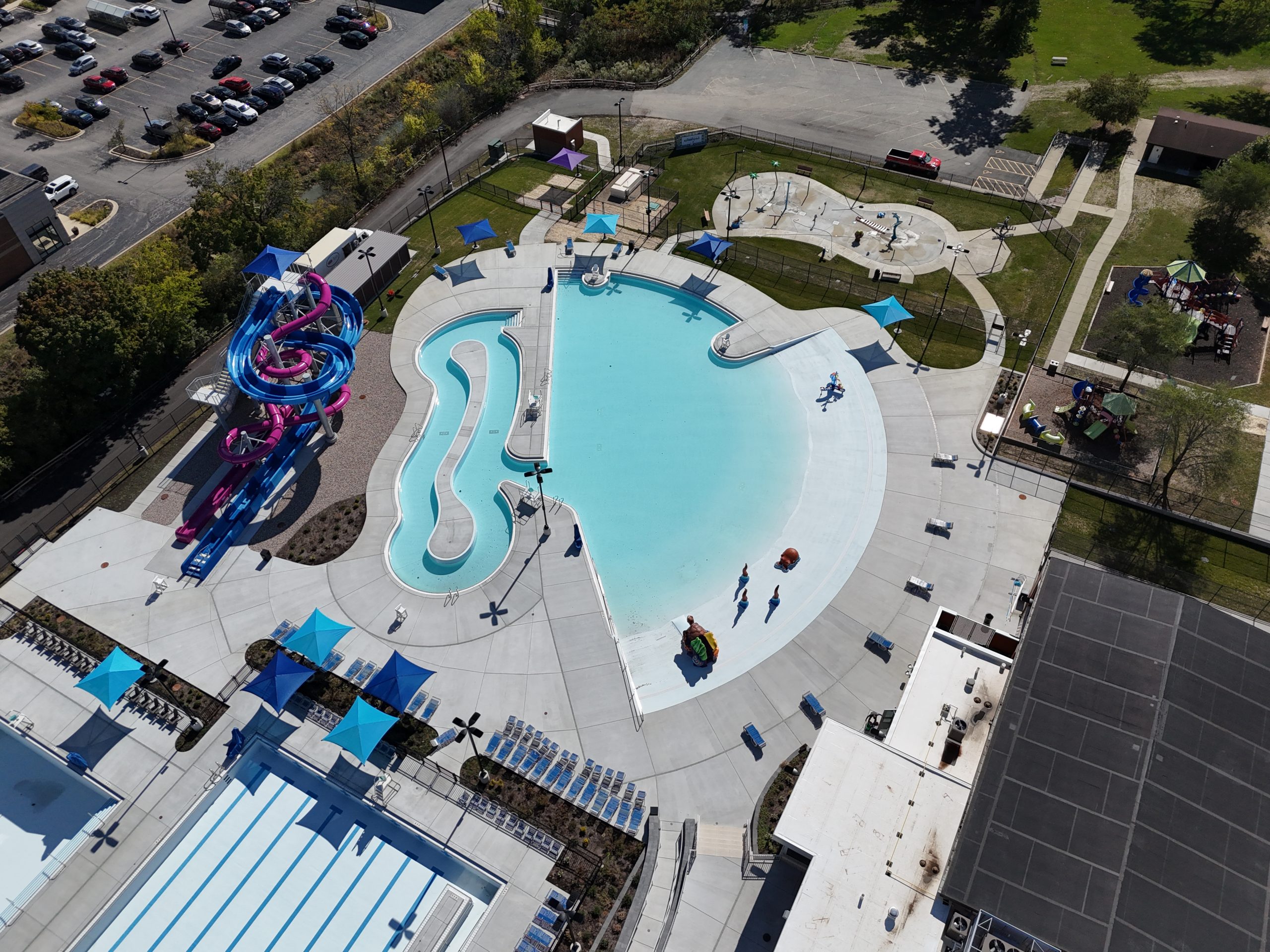 default Aerial view of a modern splash park featuring a large pool, water slides, shaded areas, and surrounding recreational facilities.