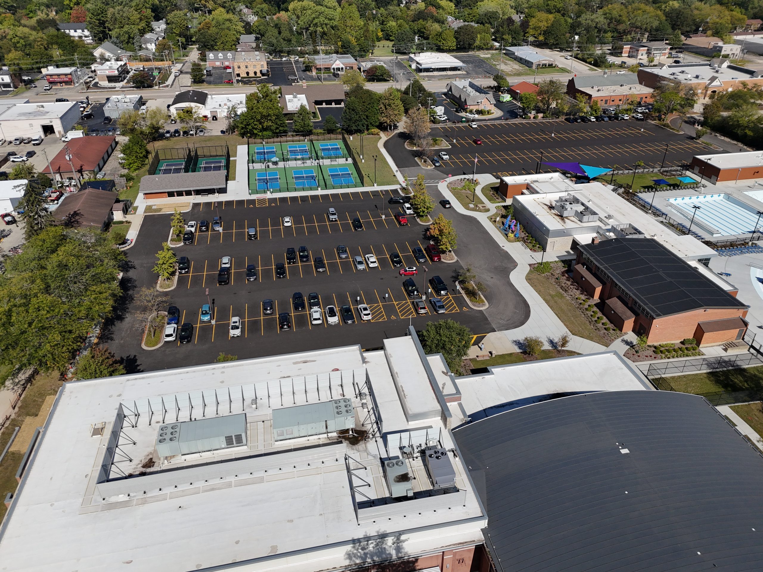 default Aerial view of a municipal recreation area featuring a parking lot, tennis courts, and modern buildings in a suburban setting.