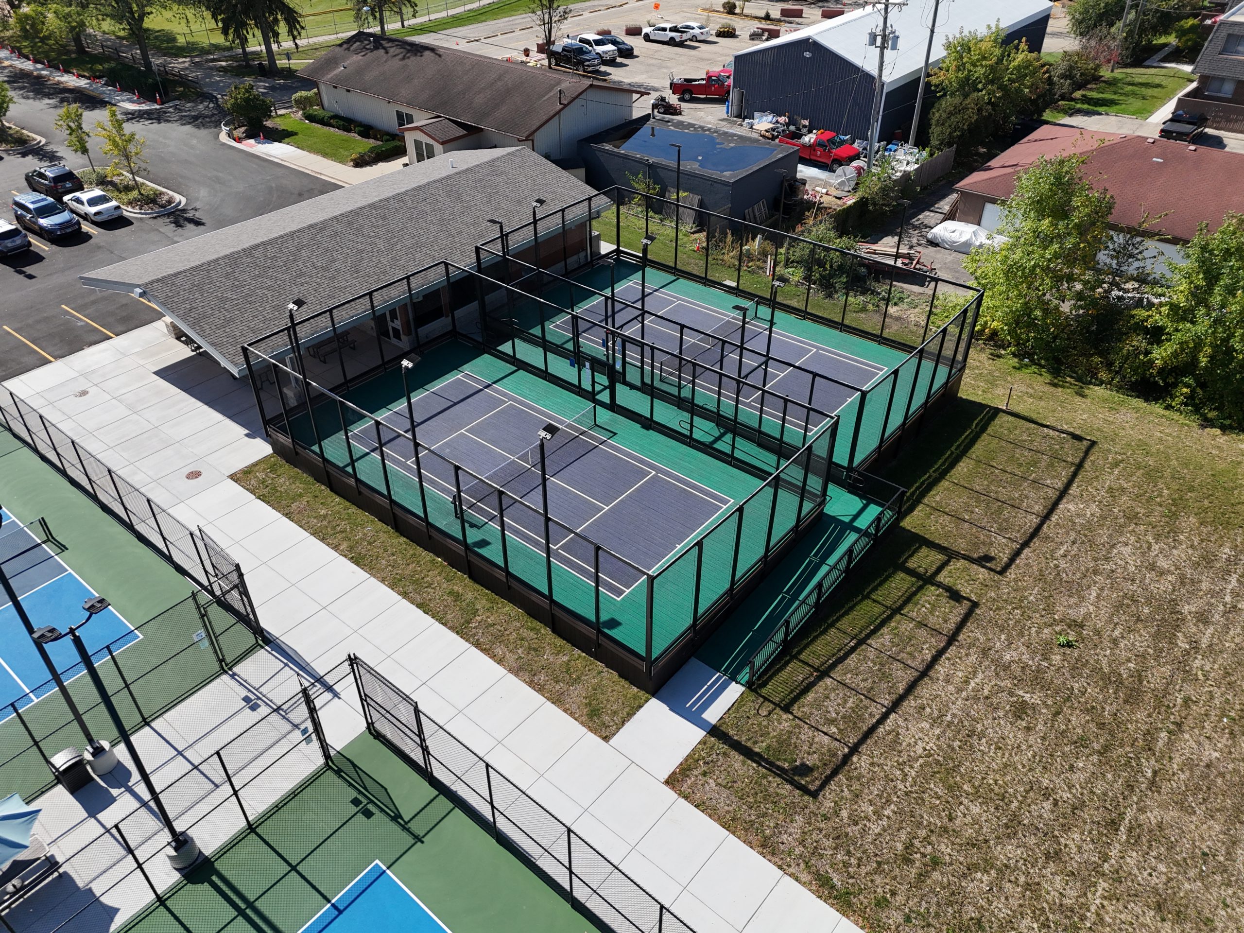 default Aerial view of a multi-court outdoor sports facility featuring pickleball and tennis courts, surrounded by landscaped areas and buildings.