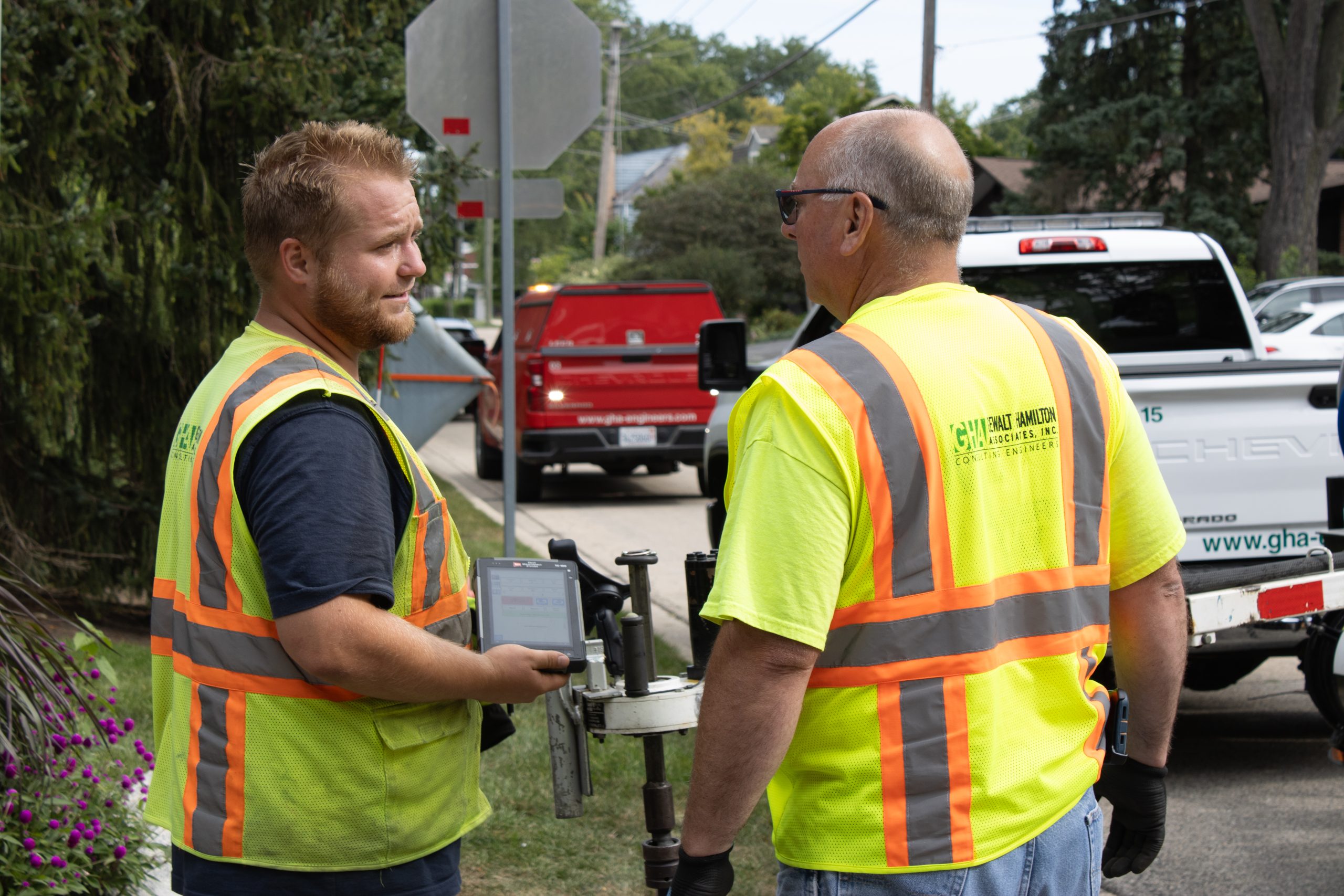 IMG_4728 Two civil engineers in safety vests discuss project details while holding surveying equipment near a vehicle on a municipal street.