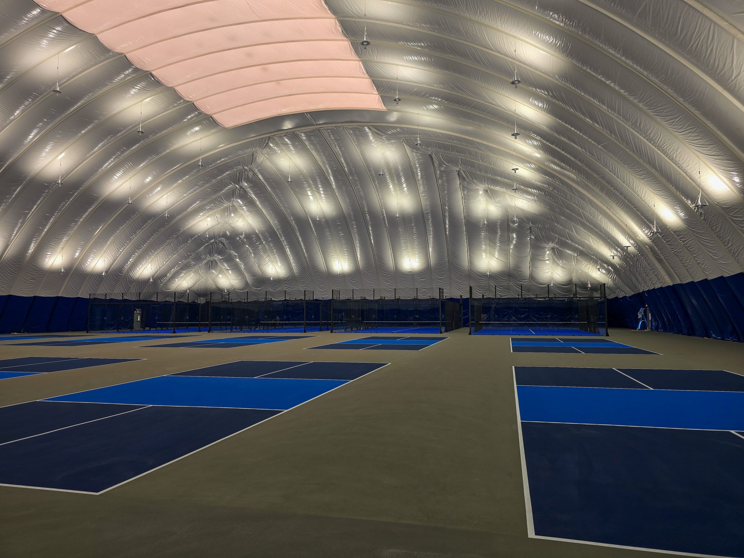 20250806_100157-2 Interior of an inflatable tennis dome showcasing multiple blue courts with bright overhead lighting.