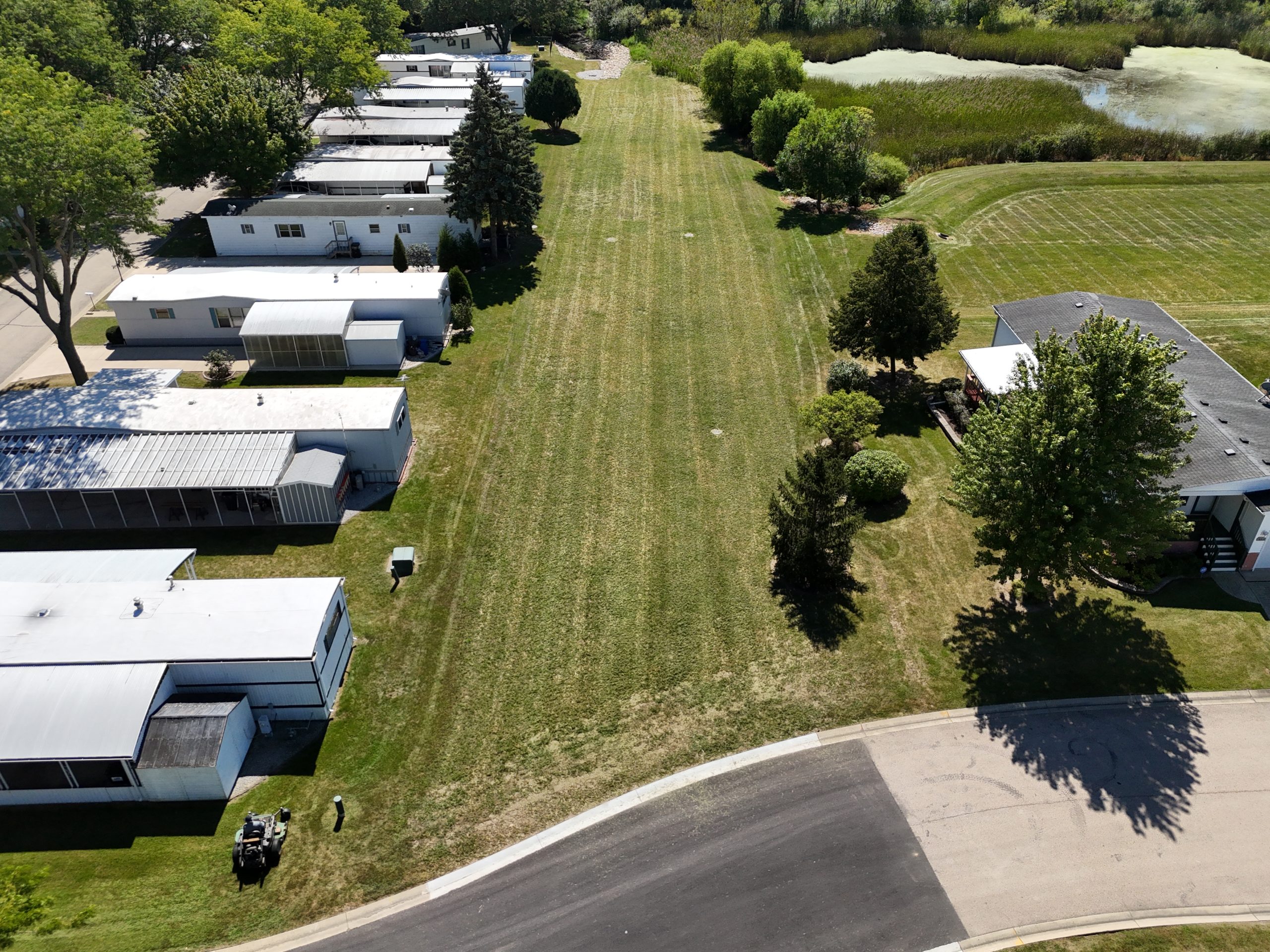 default Aerial view of a well-maintained green space beside a row of mobile homes, featuring trees and a nearby pond.