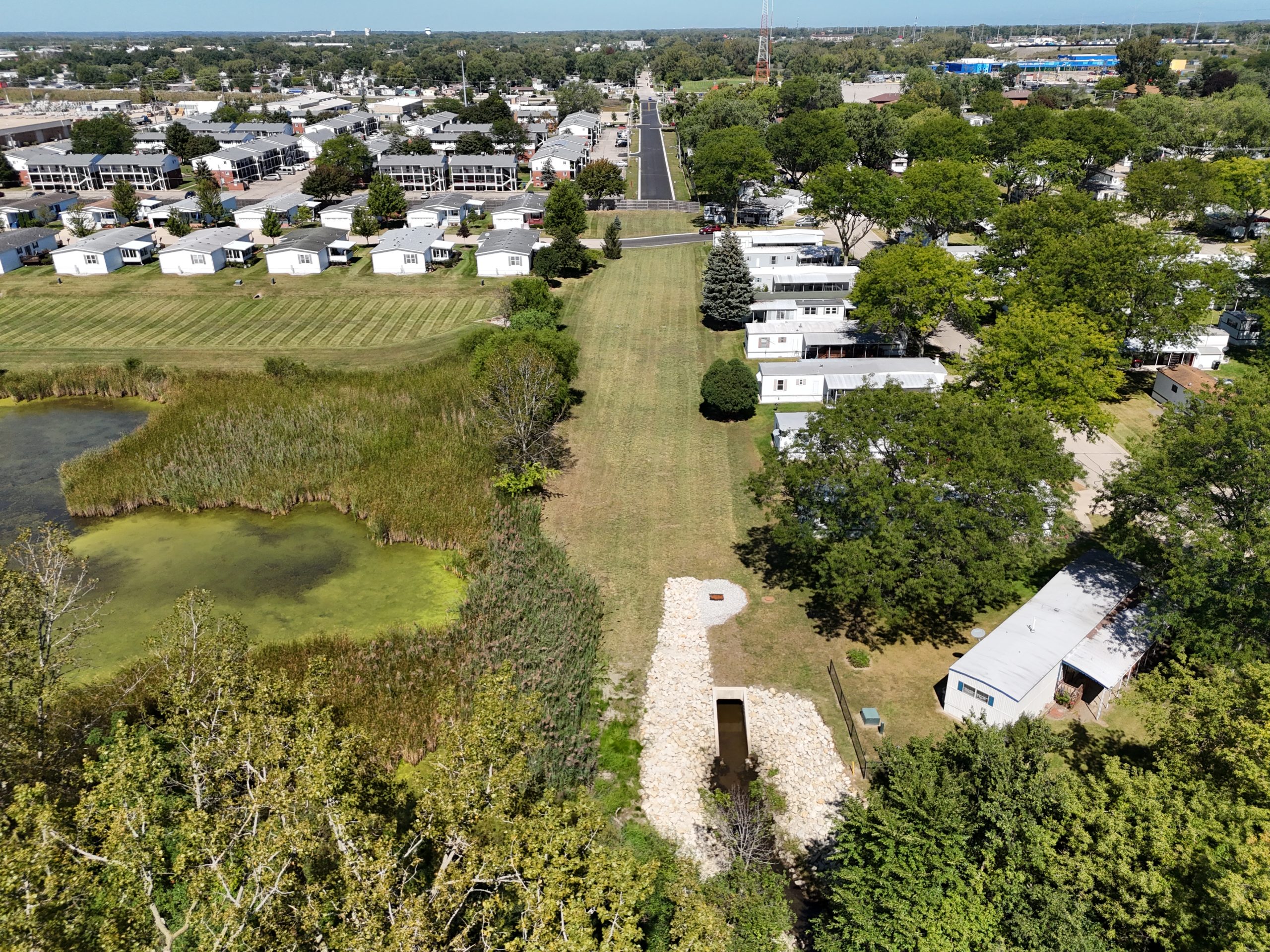 default Aerial view of a park area featuring a pond, grassy spaces, and mobile homes, with municipal roads and greenery in the background.